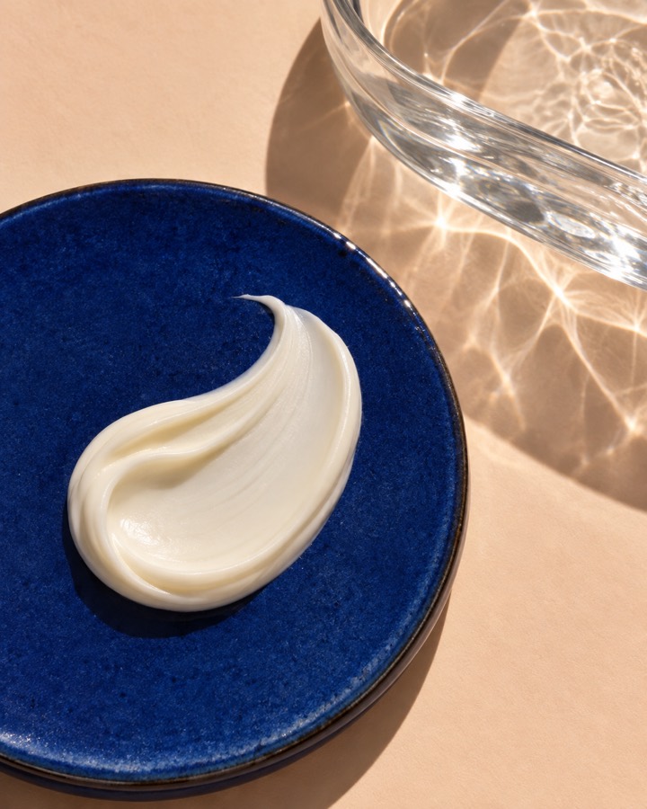 A swirl of skincare cream on a blue tile beside a clear glass dish